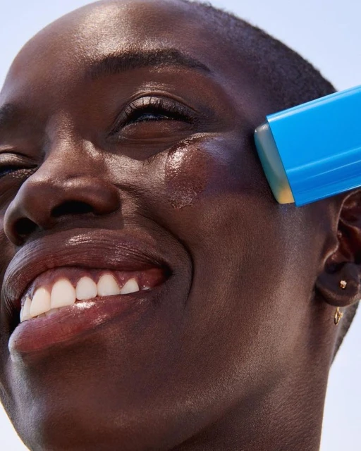 Close-up of a Black woman applying a blue sunscreen stick to her cheek, smiling with white teeth visible against a light background.