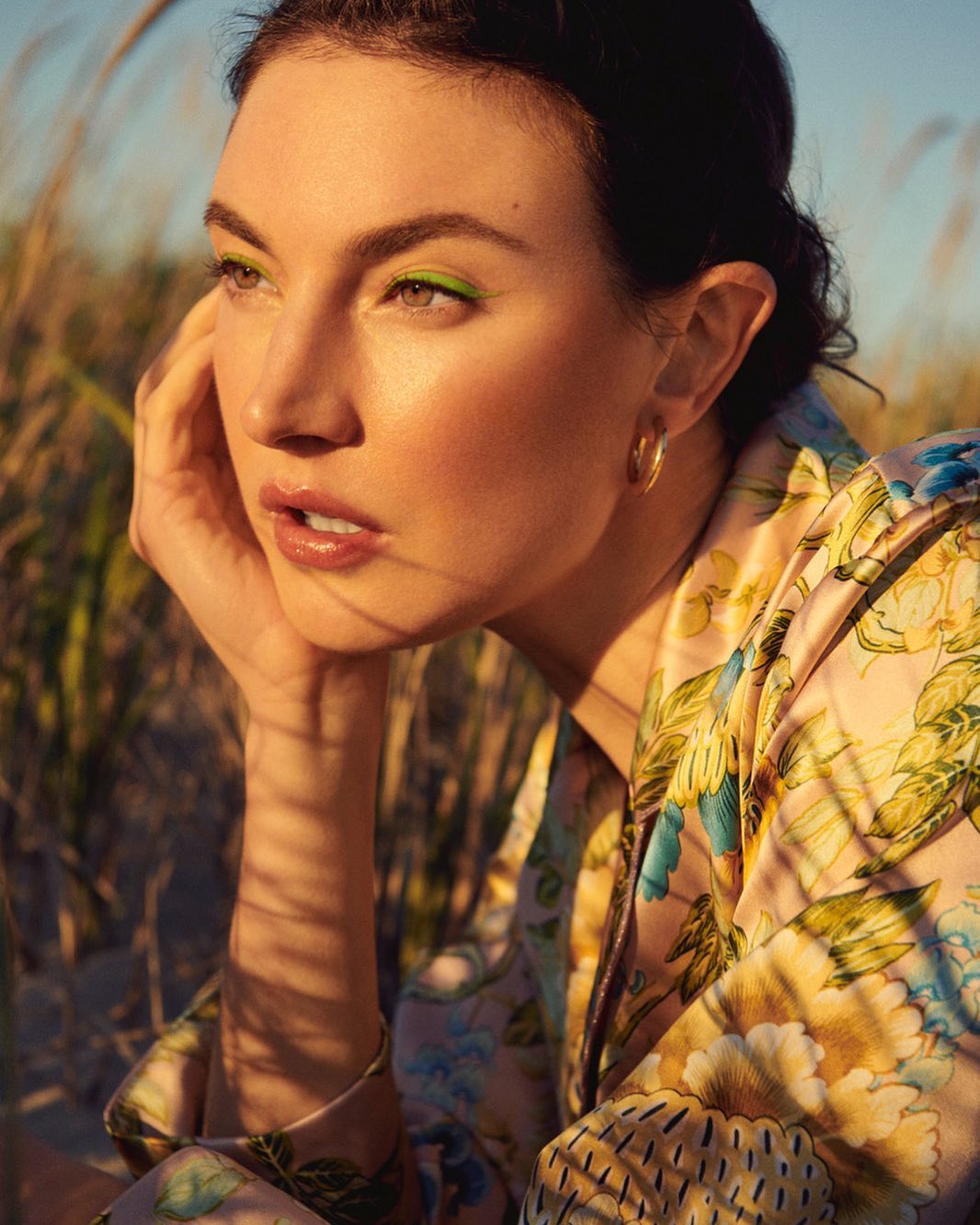 Portrait of a woman with bright green eyeliner outdoors in floral shirt looking to the side with grasses and blue sky in the background.