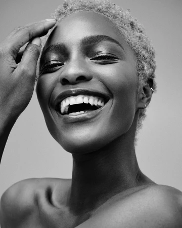 Close-up portrait of a smiling Black woman with short blonde hair touching her forehead against a plain background