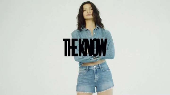 Brunette woman in light-blue denim shirt and shorts standing against a white background with the text "The Know" superimposed.