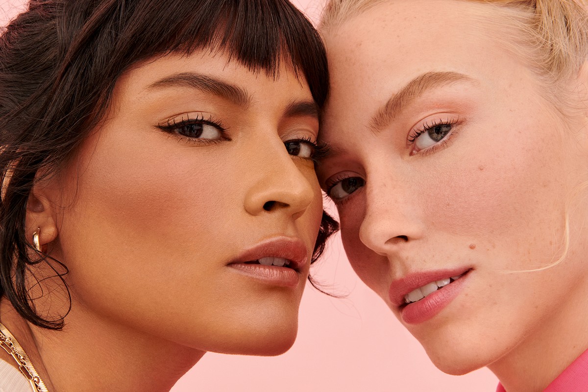 Close-up studio shot of two adult women with fair and medium complexions, showcasing makeup and gold earrings against a pink backdrop.