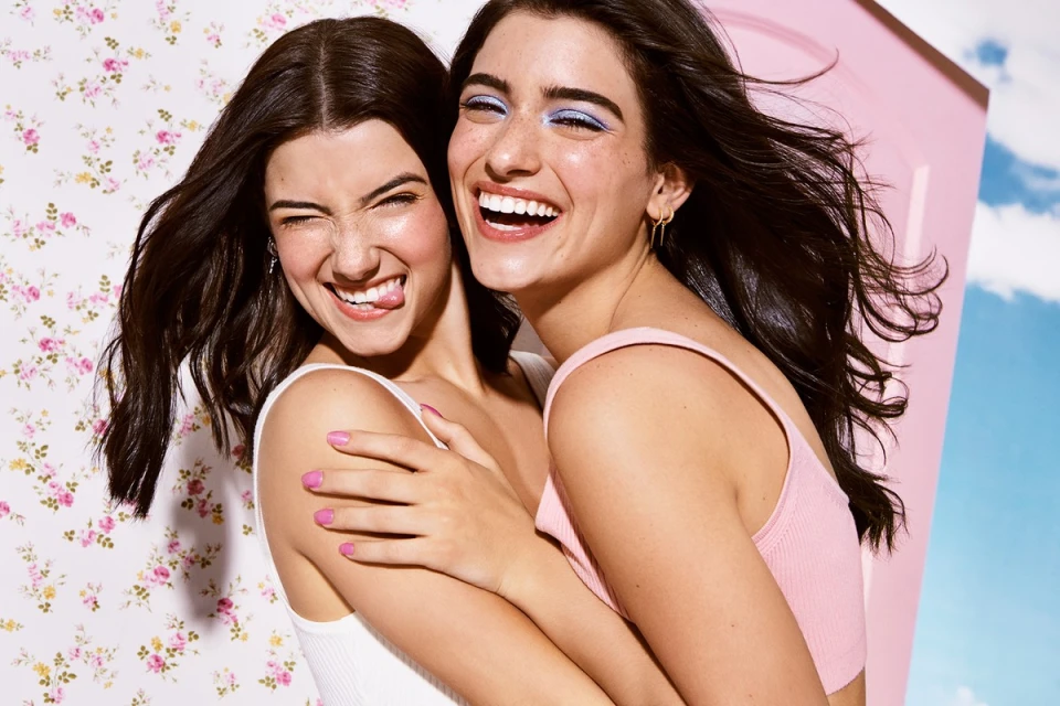 Two young women pose closely, smiling and laughing; one playfully sticks out her tongue, with a floral wall and sky backdrop.