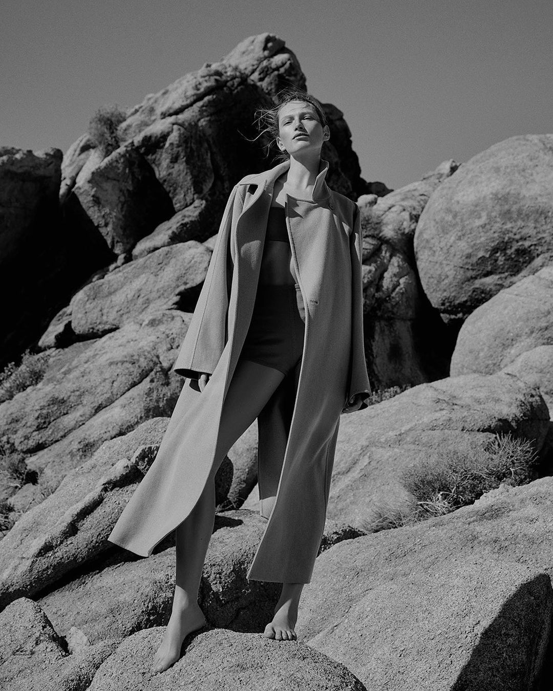 Black and white portrait of a woman standing barefoot among large rock formations wearing a long coat