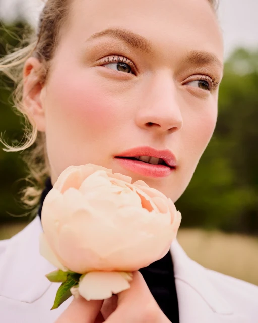 Close-up shot of a fair-skinned woman holding a light pink peony against an out-of-focus green background, wearing a black top and white jacket.