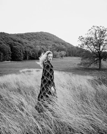 Monochrome portrait of a woman with long wavy hair standing in a grassy field wearing a plaid coat