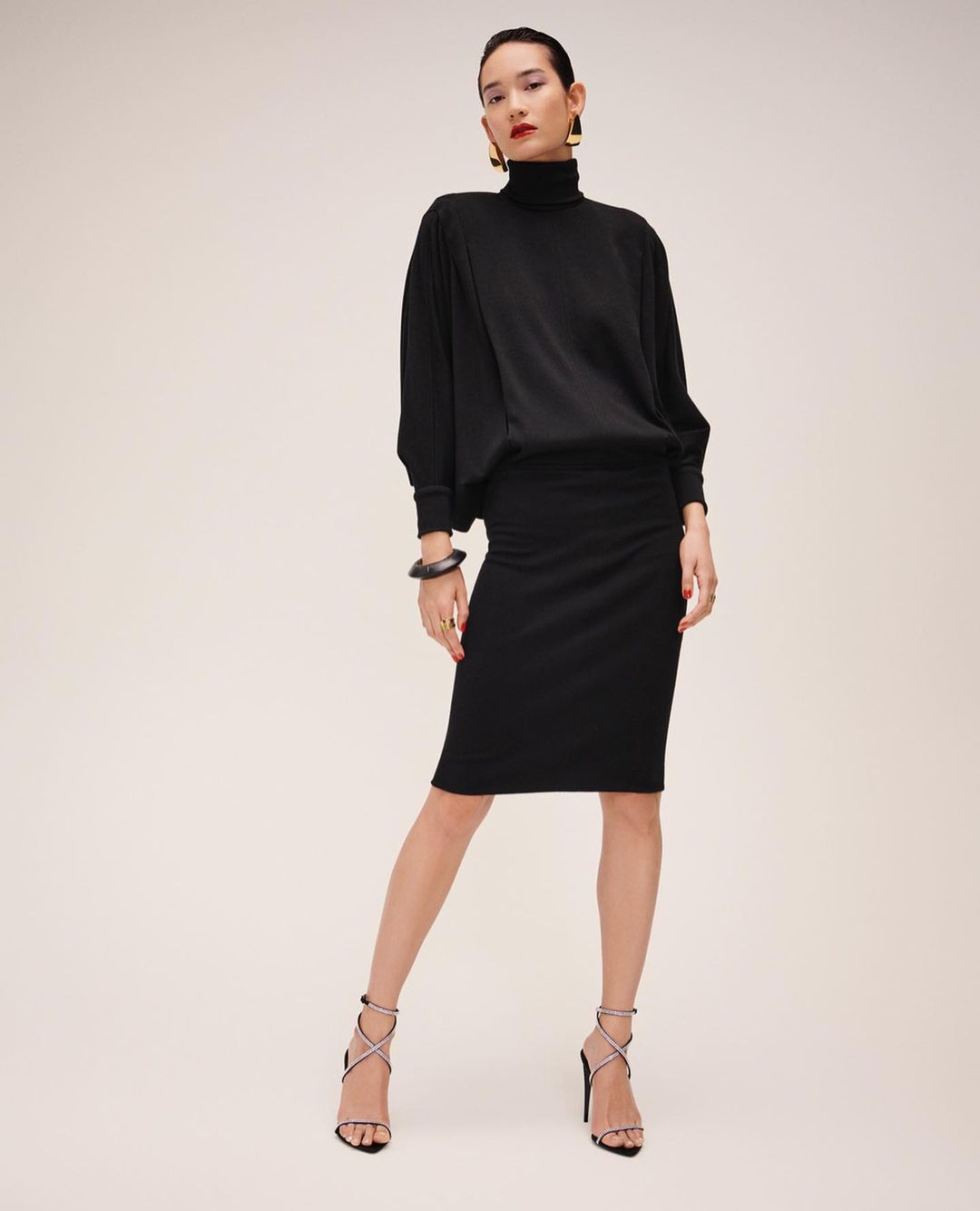 Adult female fashion model posing in a black turtleneck sweater and skirt with strappy heeled sandals in a studio setting.