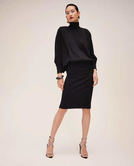 Adult female fashion model posing in a black turtleneck sweater and skirt with strappy heeled sandals in a studio setting.