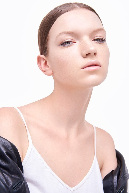 Close-up studio shot of a young adult female model in a white tank top and black jacket, gazing directly at the camera on a white background.