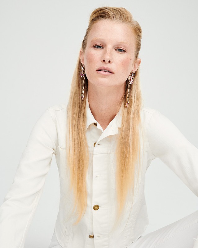 Portrait of a blonde woman wearing a white denim jacket and ornate crystal tassel earrings against a white seamless background