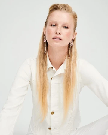 Portrait of a blonde woman wearing a white denim jacket and ornate crystal tassel earrings against a white seamless background