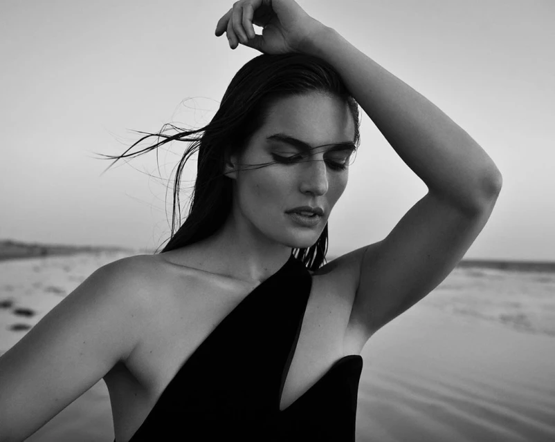 Monochrome portrait of a woman with dark hair in a black dress, posing outdoors with wind blowing through her hair against a blurred sky.