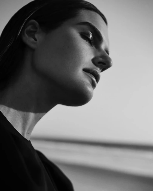 Monochrome portrait of an adult woman with sleek hair, her head tilted down, focusing on her face and neck in a neutral indoor space.