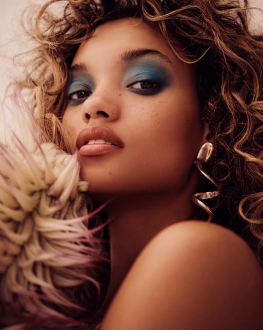 Close-up portrait of a young adult female model with bronze skin, curly brown hair, blue eyeshadow, glossy lips, and statement earrings.