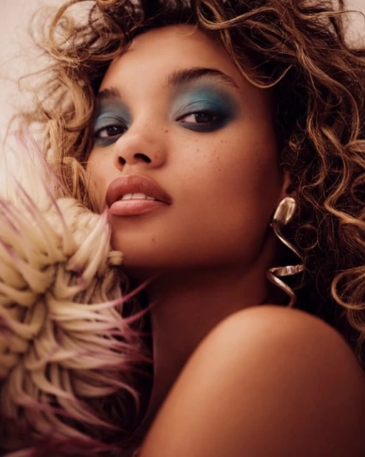Close-up portrait of a young adult female model with bronze skin, curly brown hair, blue eyeshadow, glossy lips, and statement earrings.