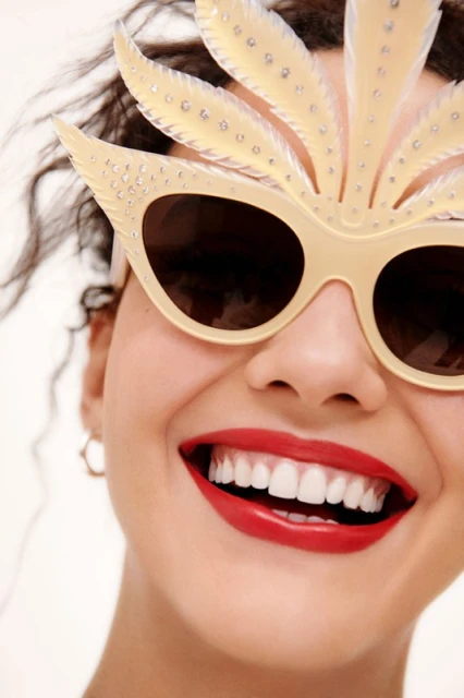 Close-up of a smiling woman with red lipstick, white teeth, and decorative sunglasses with a feathered embellishment. 