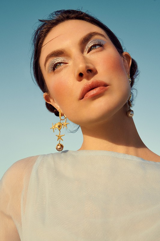 Close up portrait of a woman wearing ornate starfish drop earrings and shimmering blue eyeshadow against a clear sky.