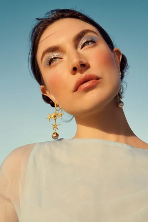 Close up portrait of a woman wearing ornate starfish drop earrings and shimmering blue eyeshadow against a clear sky.