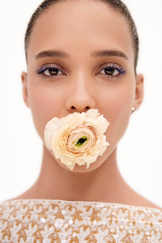 Close-up of a woman with a flower in her mouth, featuring striking blue eyelashes and minimal makeup against a neutral backdrop.