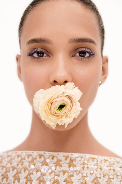 Close-up of a woman with a flower in her mouth, featuring striking blue eyelashes and minimal makeup against a neutral backdrop.