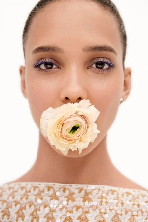 Close-up of a woman with a flower in her mouth, featuring striking blue eyelashes and minimal makeup against a neutral backdrop.