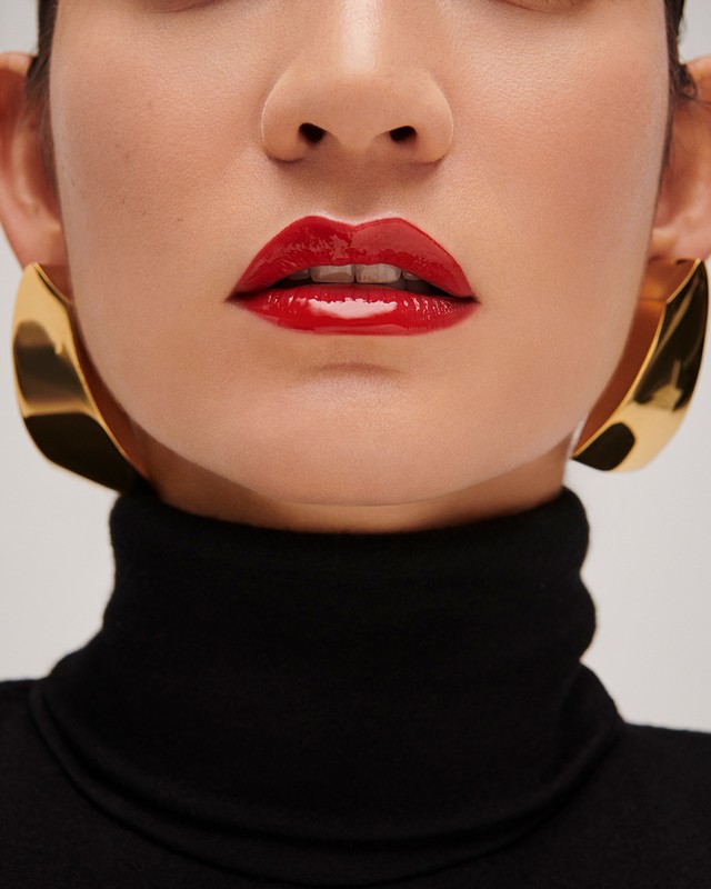Close-up beauty portrait of a woman wearing glossy red lipstick and large gold earrings with a black turtleneck against a white background.
