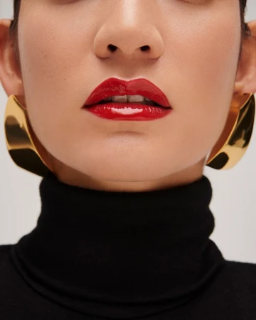 Close-up beauty portrait of a woman wearing glossy red lipstick and large gold earrings with a black turtleneck against a white background.