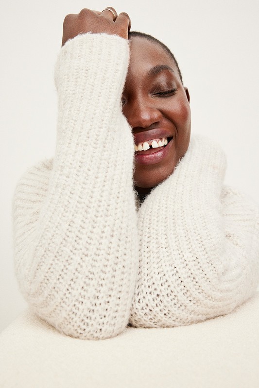 Smiling African-descent adult woman with dark hair and fair skin, wearing a knitted sweater indoors against a white background.