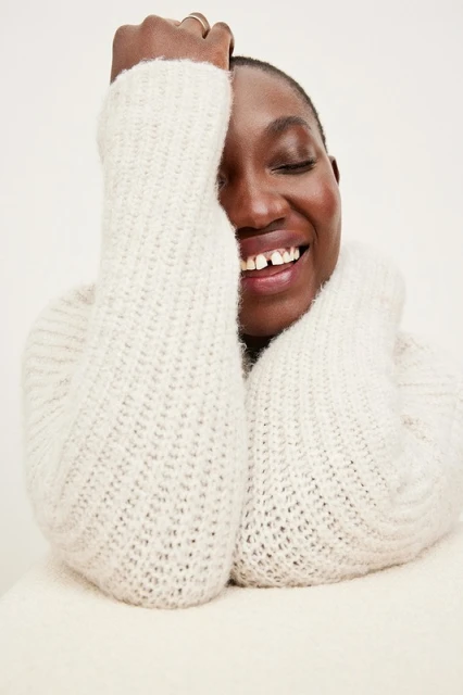 Smiling African-descent adult woman with dark hair and fair skin, wearing a knitted sweater indoors against a white background.