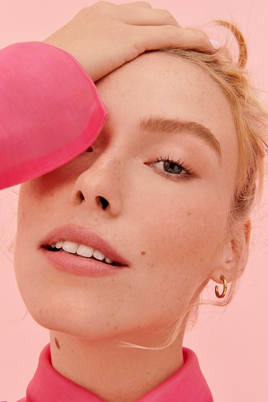 Close-up portrait of a young adult woman with light skin and blonde hair, wearing a pink shirt and gold earrings against a pink background.