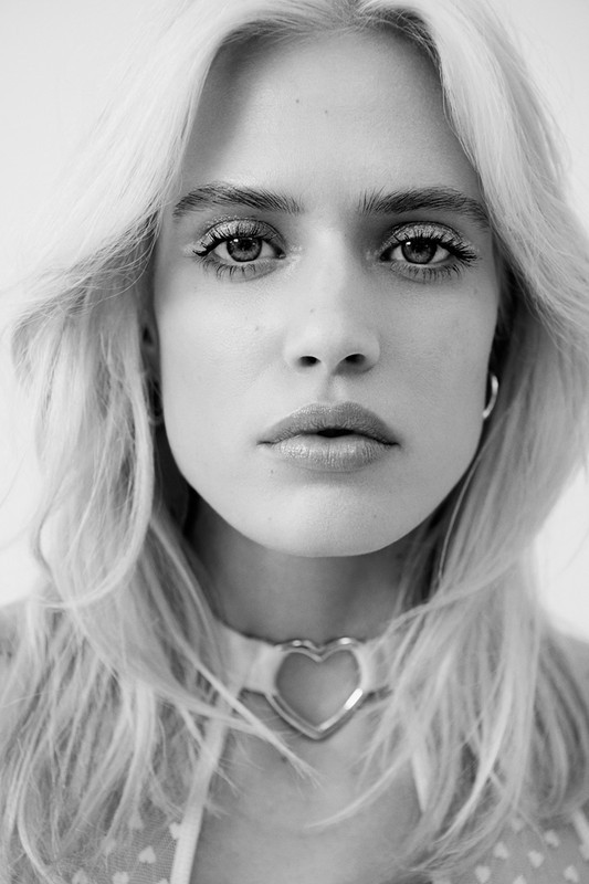 Close-up portrait of a woman with blonde hair wearing a heart choker necklace against a white background