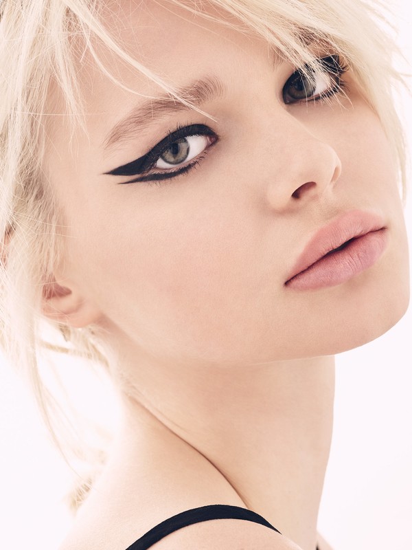 Close-up beauty portrait of a blonde woman with dramatic black winged eyeliner and pale pink lips against a plain light background