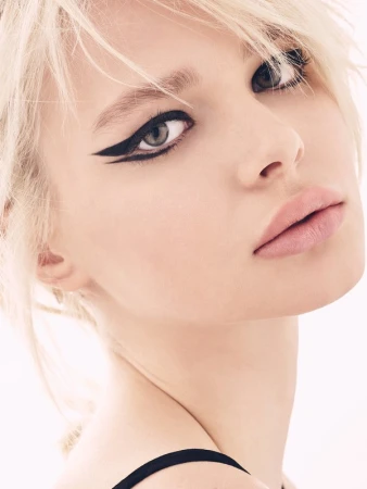 Close-up beauty portrait of a blonde woman with dramatic black winged eyeliner and pale pink lips against a plain light background