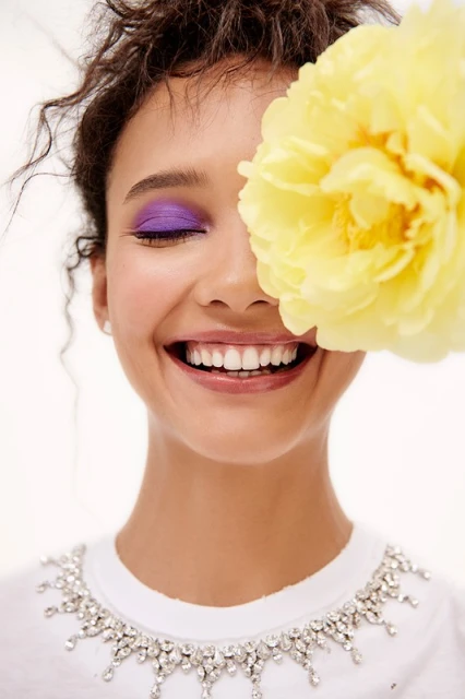 Smiling woman with purple eyeshadow and yellow flower partially covering face, wearing a white shirt with jeweled neckline in a studio.