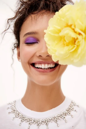 Smiling woman with purple eyeshadow and yellow flower partially covering face, wearing a white shirt with jeweled neckline in a studio.