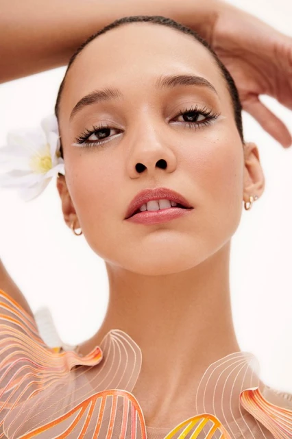 Close-up portrait of a young woman with a flower in her hair, wearing an abstract patterned top against a bright white background.