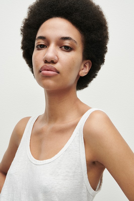 Portrait of a woman with a large afro hairstyle and nose ring wearing a white tank top against a plain background