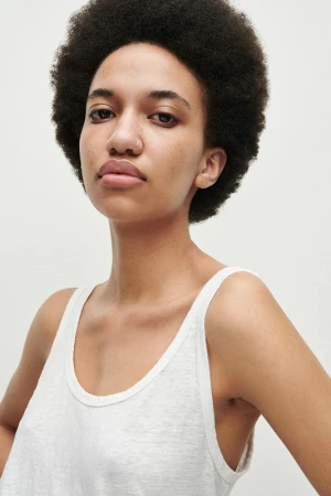 Portrait of a woman with a large afro hairstyle and nose ring wearing a white tank top against a plain background