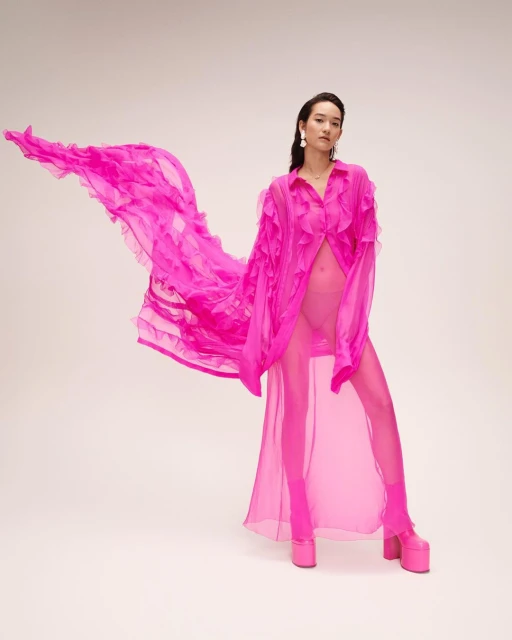 A woman in a semi-sheer fuchsia dress and platform heels poses against a neutral background, with flowing ruffles creating movement.