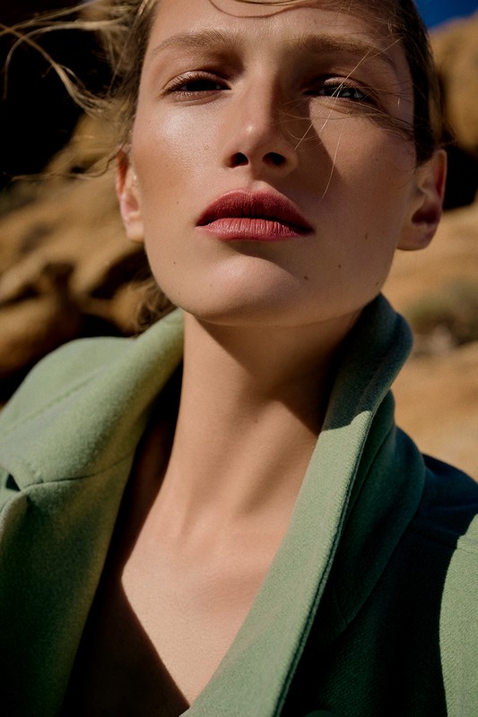 Close-up of a model in a light green coat posing in a desert environment with wind blowing through her hair, bathed in warm sunlight.