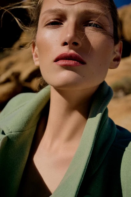 Close-up of a model in a light green coat posing in a desert environment with wind blowing through her hair, bathed in warm sunlight.