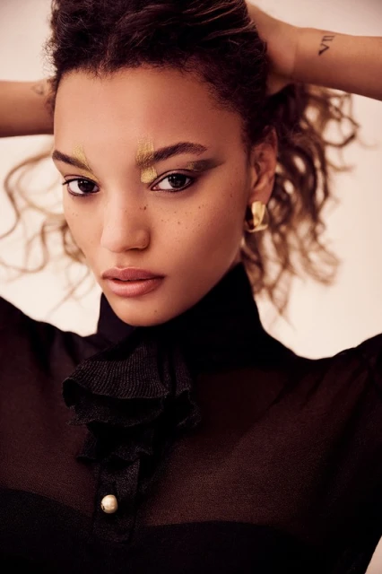 Portrait of a young woman with dramatic gold and black eye makeup, hoop earrings, and a sheer black high-necked top in a studio.