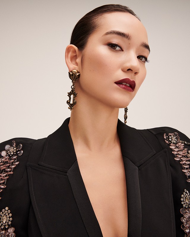 Close-up portrait of an adult female model wearing a black blazer with jeweled embellishments and antique gold statement earrings against a neutral background.