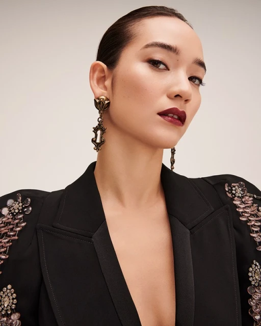 Close-up portrait of an adult female model wearing a black blazer with jeweled embellishments and antique gold statement earrings against a neutral background.