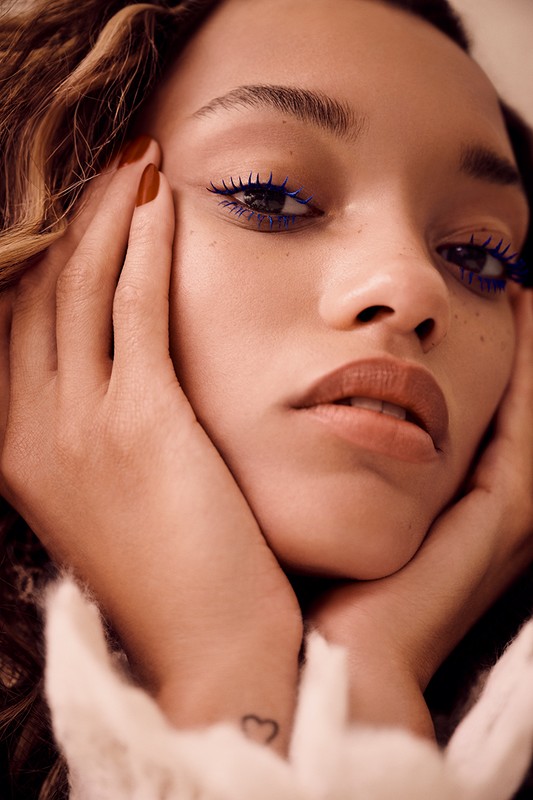 Close-up portrait of a young adult female with neutral expression, blue mascara, and brown hair, with hands framing her face.