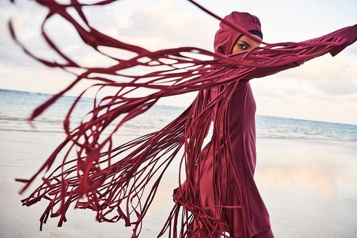 Woman in hooded swimwear on beach with red fringe detail blowing in wind, ocean and sky in background. Fashion, swimwear.