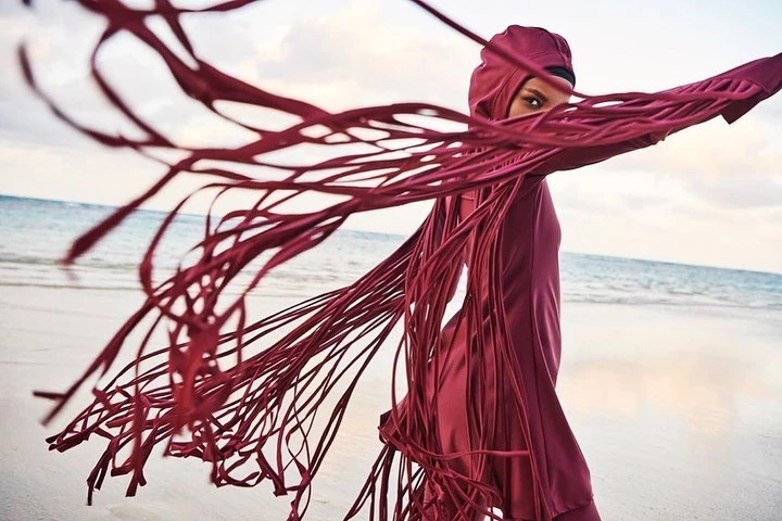 Woman in hooded swimwear on beach with red fringe detail blowing in wind, ocean and sky in background. Fashion, swimwear.