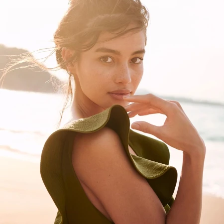 Close-up portrait of a woman wearing an olive green textured top against a bright beach background with sun flare.