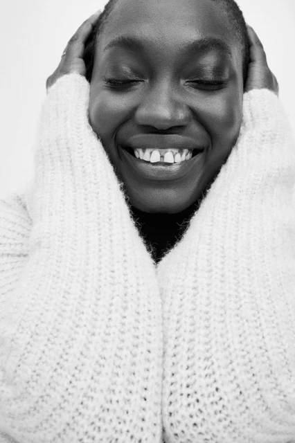 Close-up black and white portrait of a smiling African woman with closed eyes wearing a textured sweater with hands near her face.