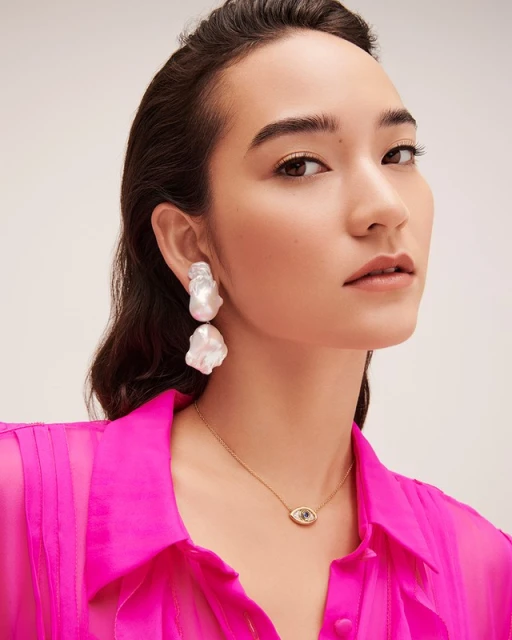 Model wearing baroque pearl drop earrings and necklace, closeup studio portrait on white backdrop, wearing bright pink blouse.