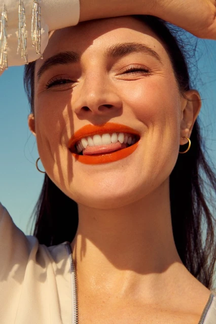 Smiling woman with orange lipstick poses outdoors under bright sunlight with a blue sky background, displaying a playful, happy expression.